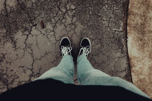 Person wearing casual jeans and sneakers standing on cracked pavement.