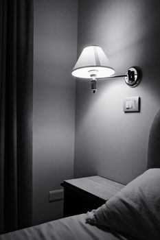 A minimalistic black and white image of a wall-mounted lamp beside a bed, casting a soft glow.