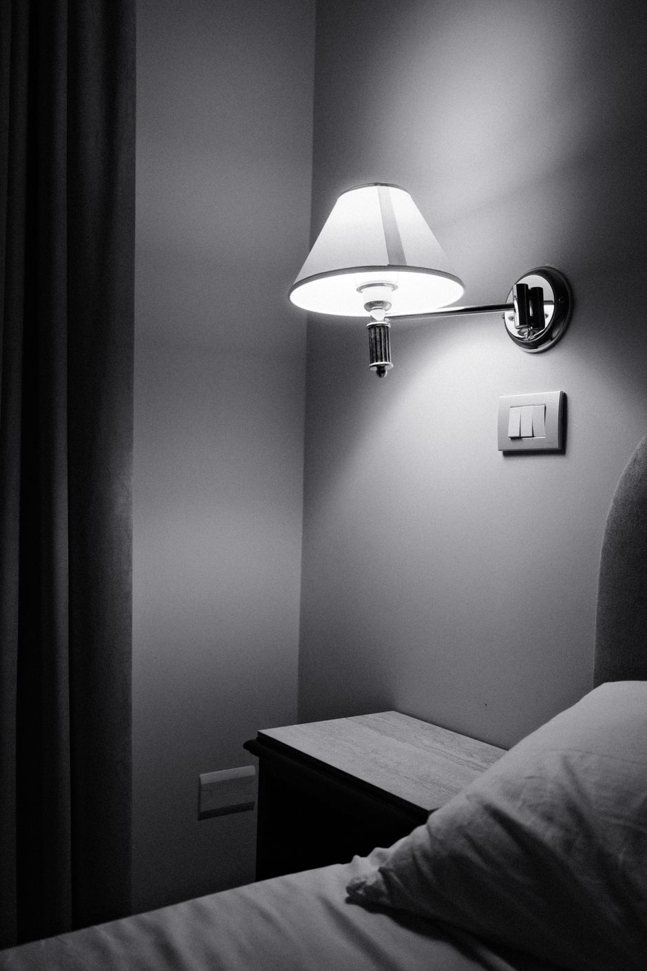 A minimalistic black and white image of a wall-mounted lamp beside a bed, casting a soft glow.