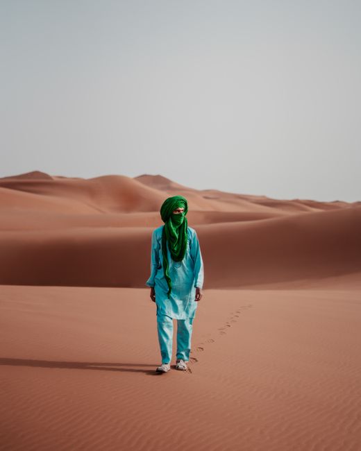 A person dressed in blue and green stands amidst the vast dunes of the Sahara Desert in Morocco.