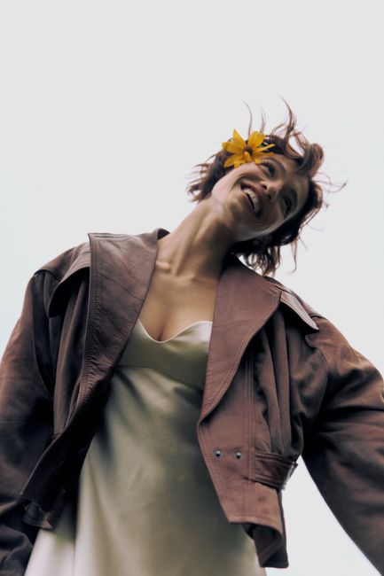 Woman outdoors with a bright smile, wearing a leather jacket and flower in hair, showcasing carefree fashion.