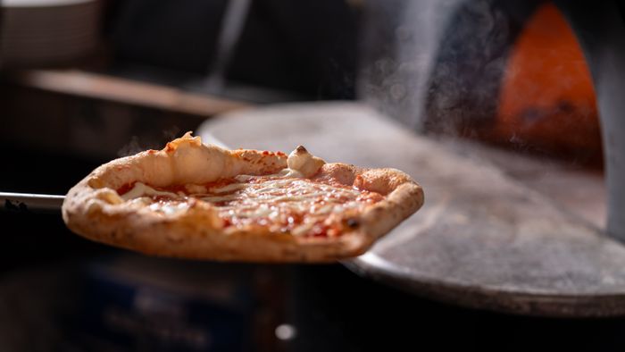 Pizza entrando al horno de leña sobre una pala, con el fondo iluminado por el fuego