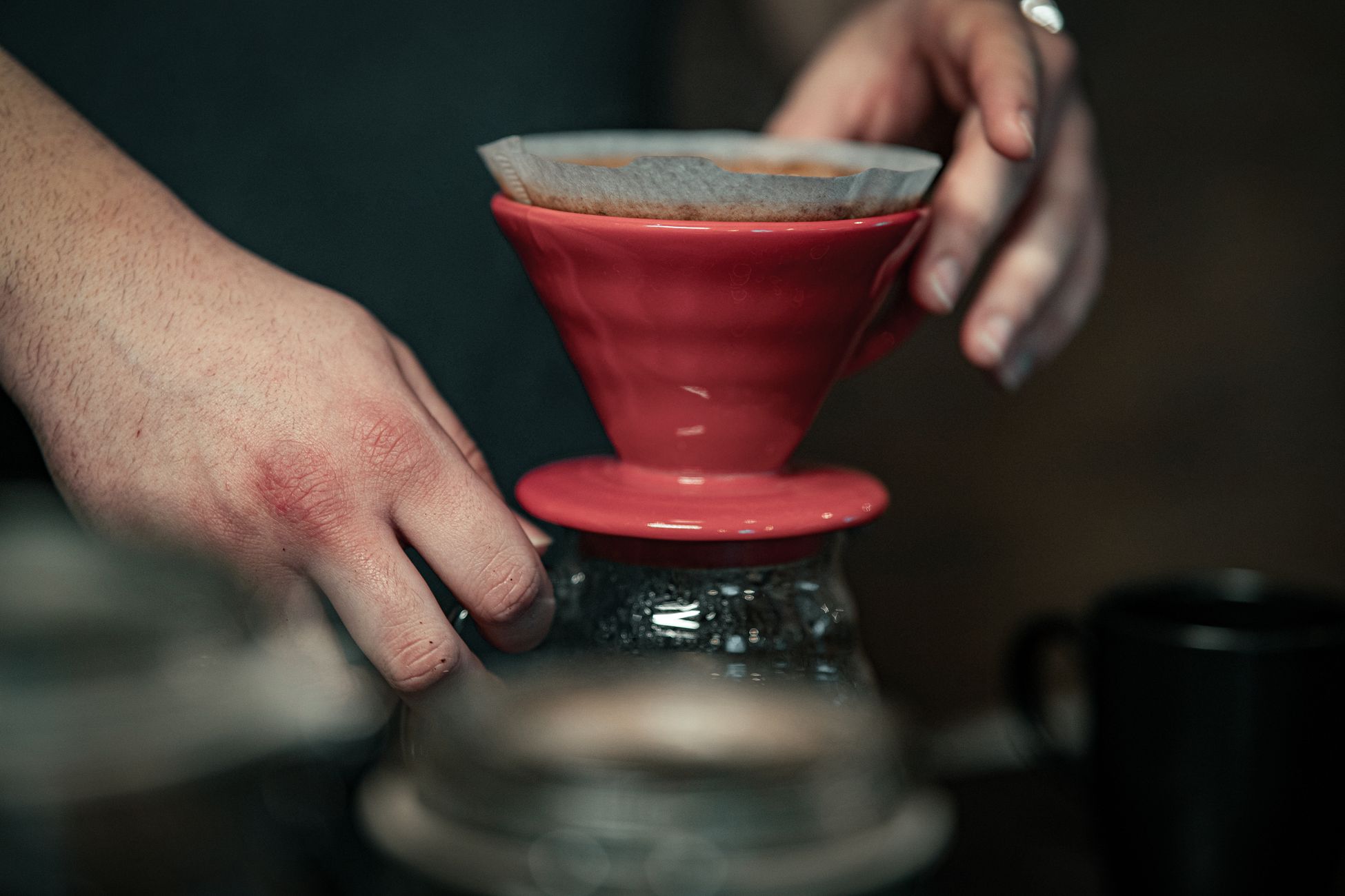 Hands preparing coffee using a red pour-over device and filter paper.