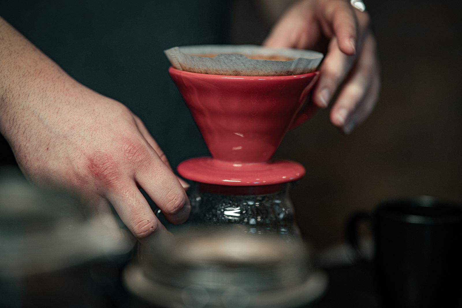 Hands steadying a red ceramic pour-over dripper with paper filter on glass carafe