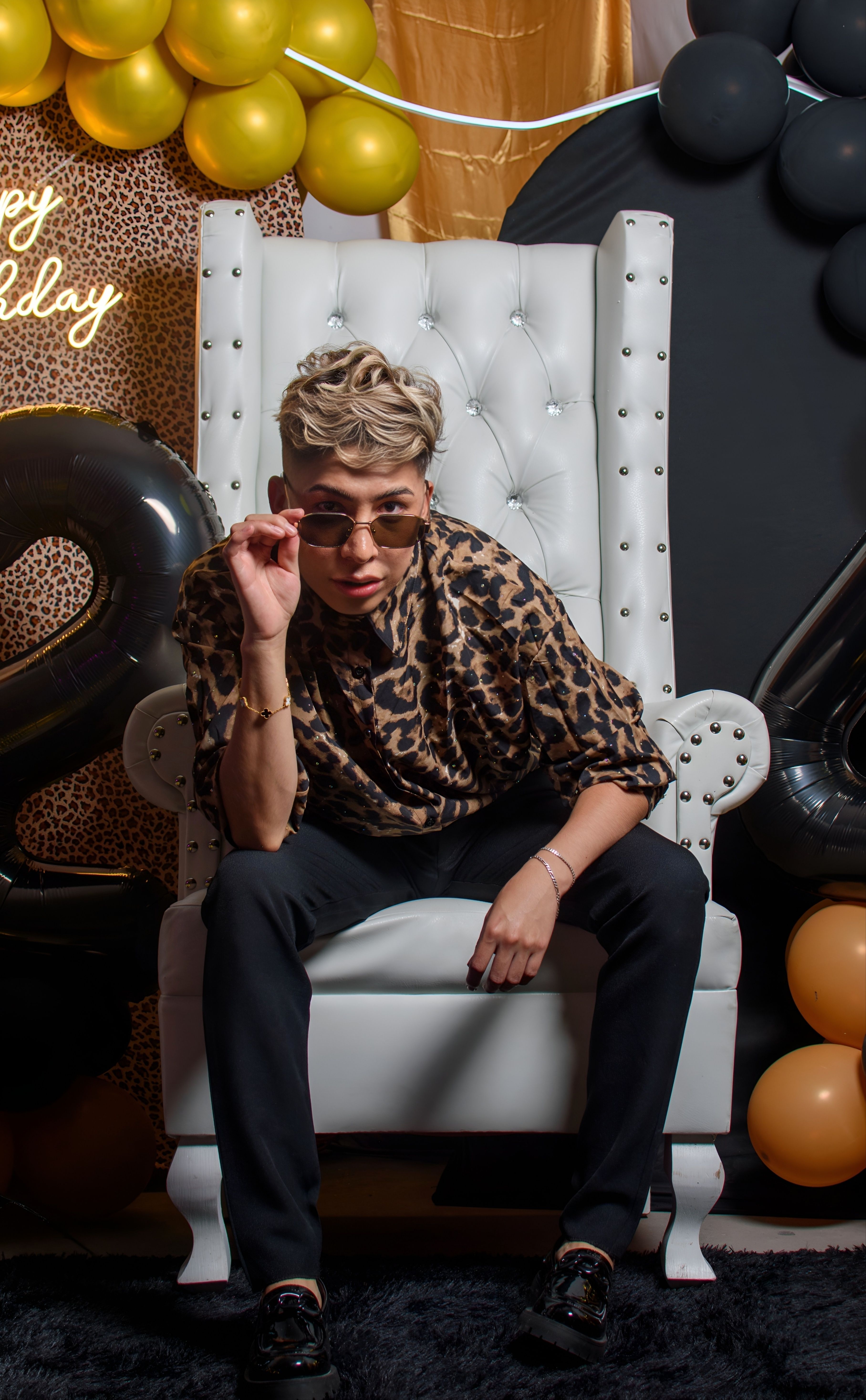 An adult in chic leopard print outfit poses on a white throne, surrounded by balloons at a birthday party in Ciudad de México.