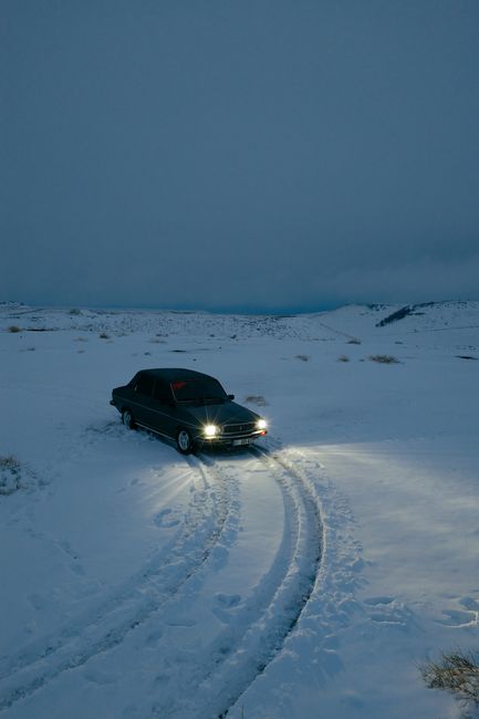 A lone car with headlights on navigates through snow-covered terrain under a twilight sky in Adıyaman, Türkiye.