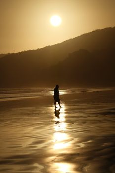 A serene silhouette of a person walking on a sunlit beach with mountains in the background.
