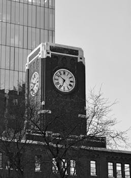 Black and white image of a classic clock tower against a modern glass building in an urban setting.