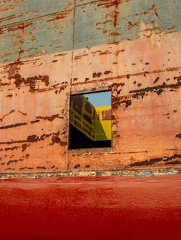 Detailed view of a rusty metal wall with a window reflecting a colorful structure.