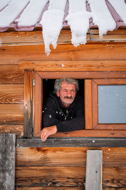 Elderly man in black sweater looks out snowy cabin window. Winter scene, rustic warmth.