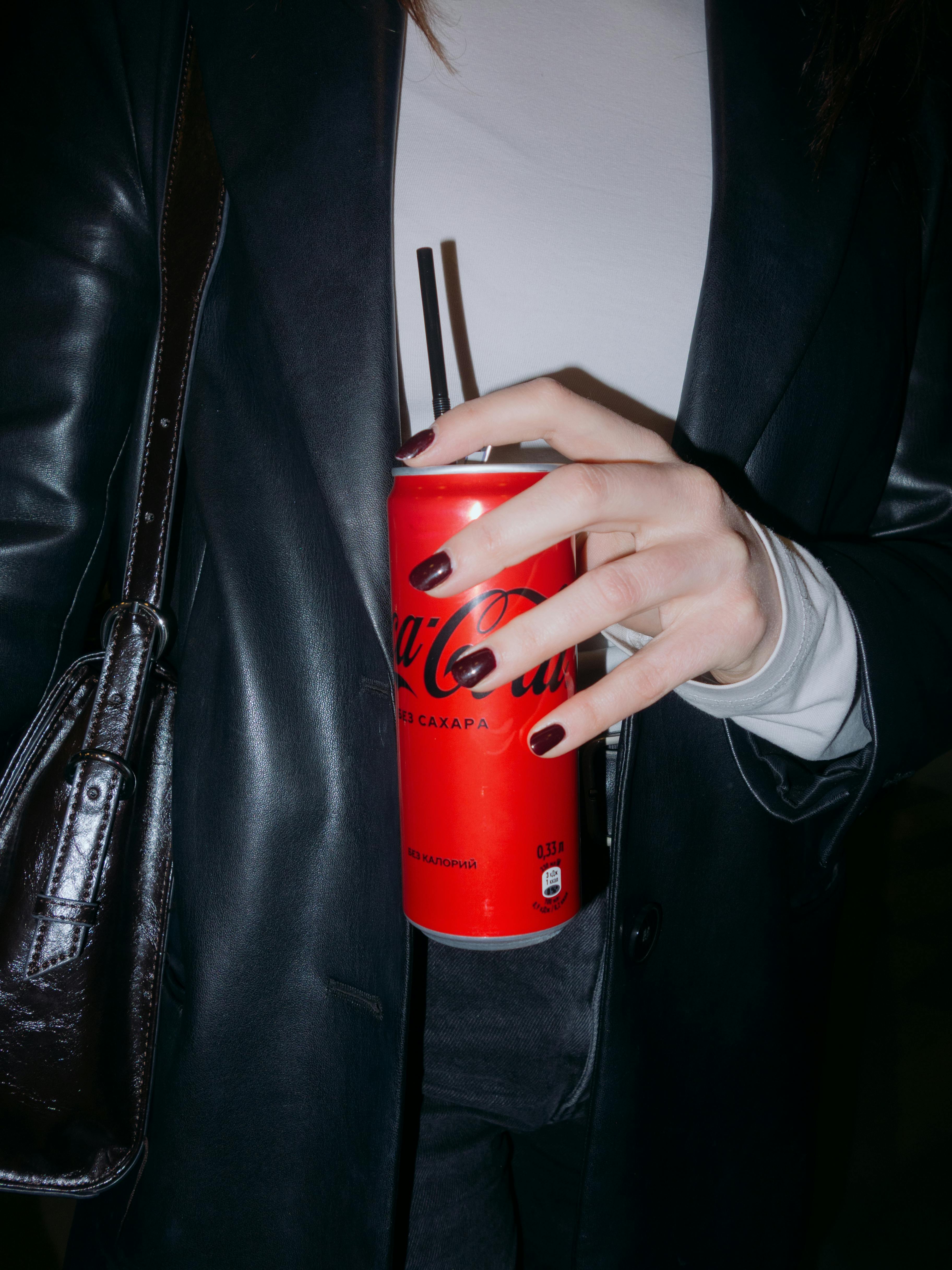 Close-up of a hand holding a red soda can with black straws, against a dark outfit background.