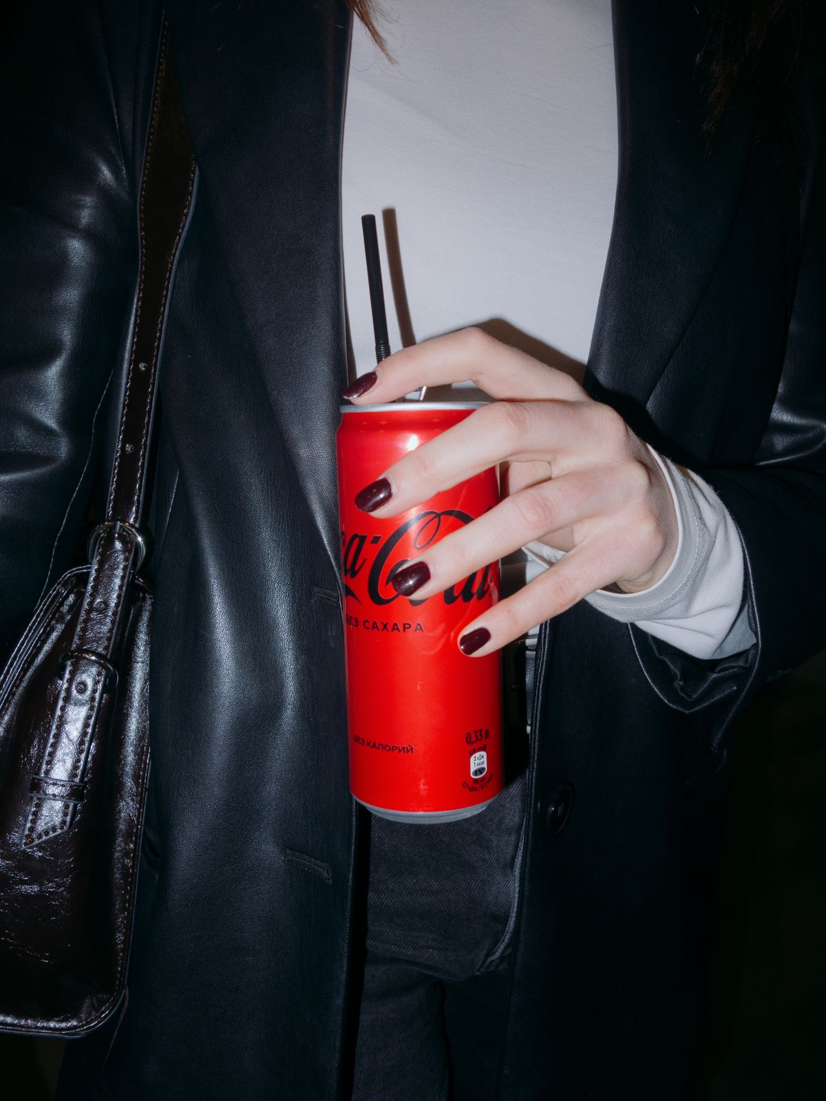 Close-up of a hand holding a red soda can with black straws, against a dark outfit background.