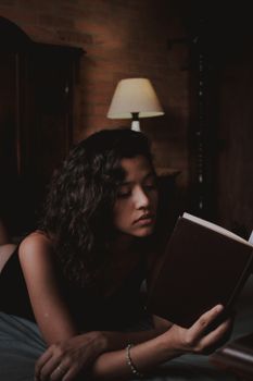 A thoughtful woman reading a book indoors with warm, moody lighting, creating a serene atmosphere.