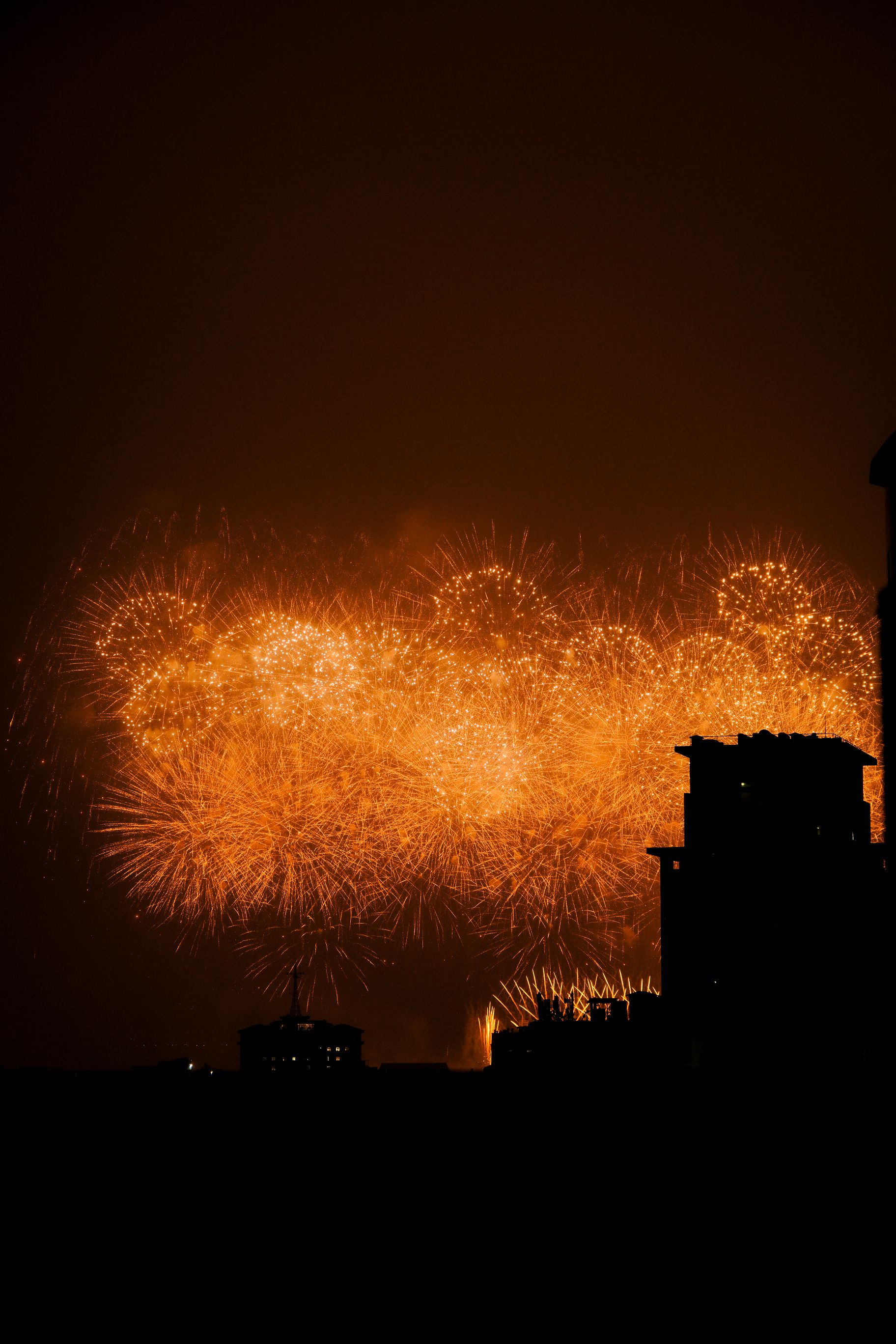 Dazzling orange fireworks illuminate the night sky, silhouetting city buildings in a captivating display.