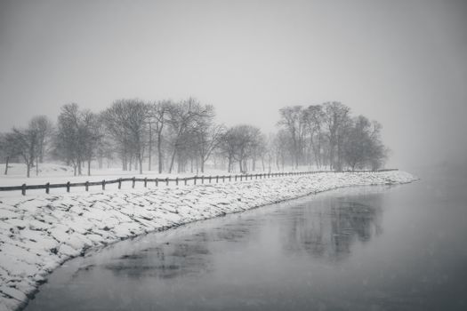 Serene winter scene of a snow-covered riverbank with barren trees reflecting in the water.