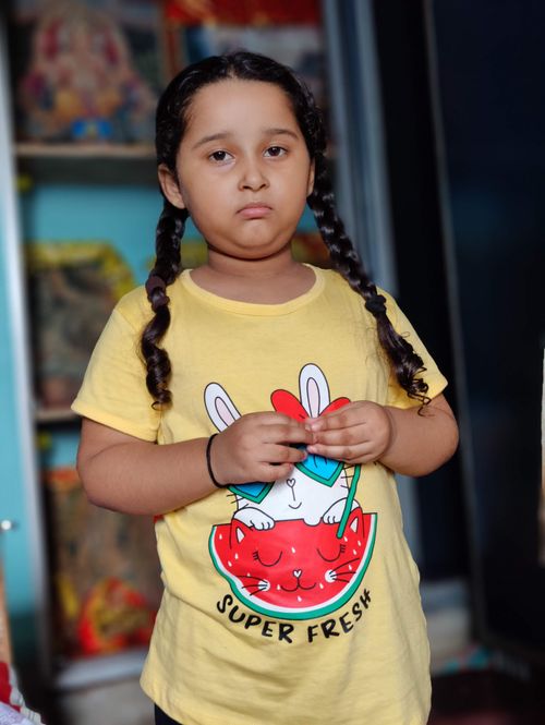 Free A young girl with braided hair wearing a yellow t-shirt with a playful design, standing indoors. Stock Photo