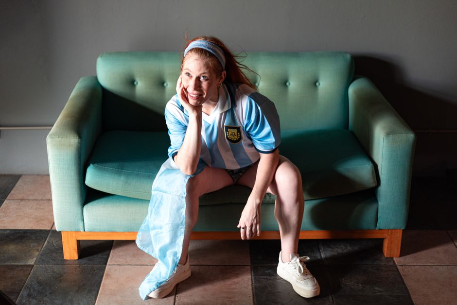 A smiling woman sitting on a teal sofa wearing a blue and white Argentina football jersey indoors.