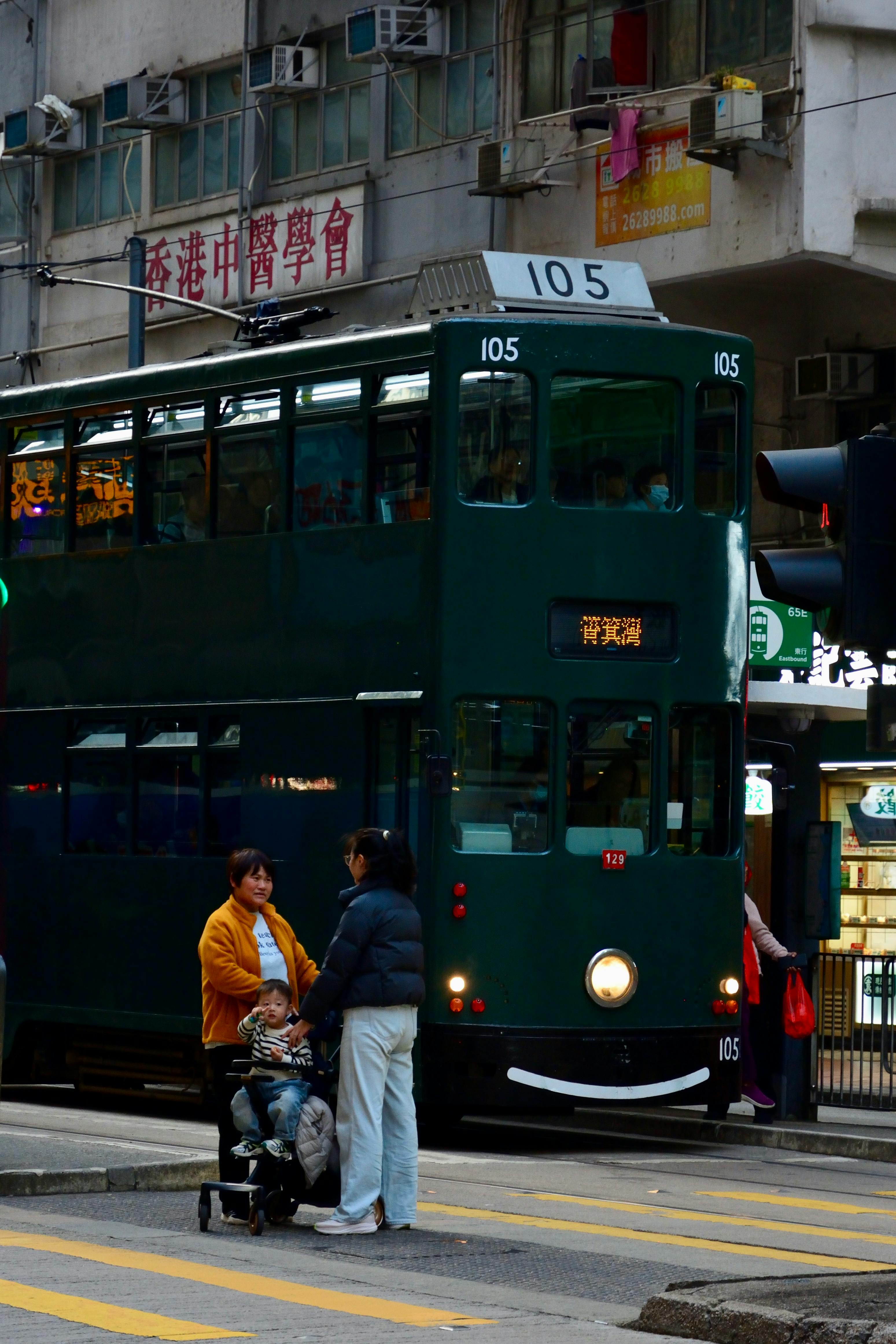 A green tram passes through a busy street in Hong Kong with pedestrians nearby.