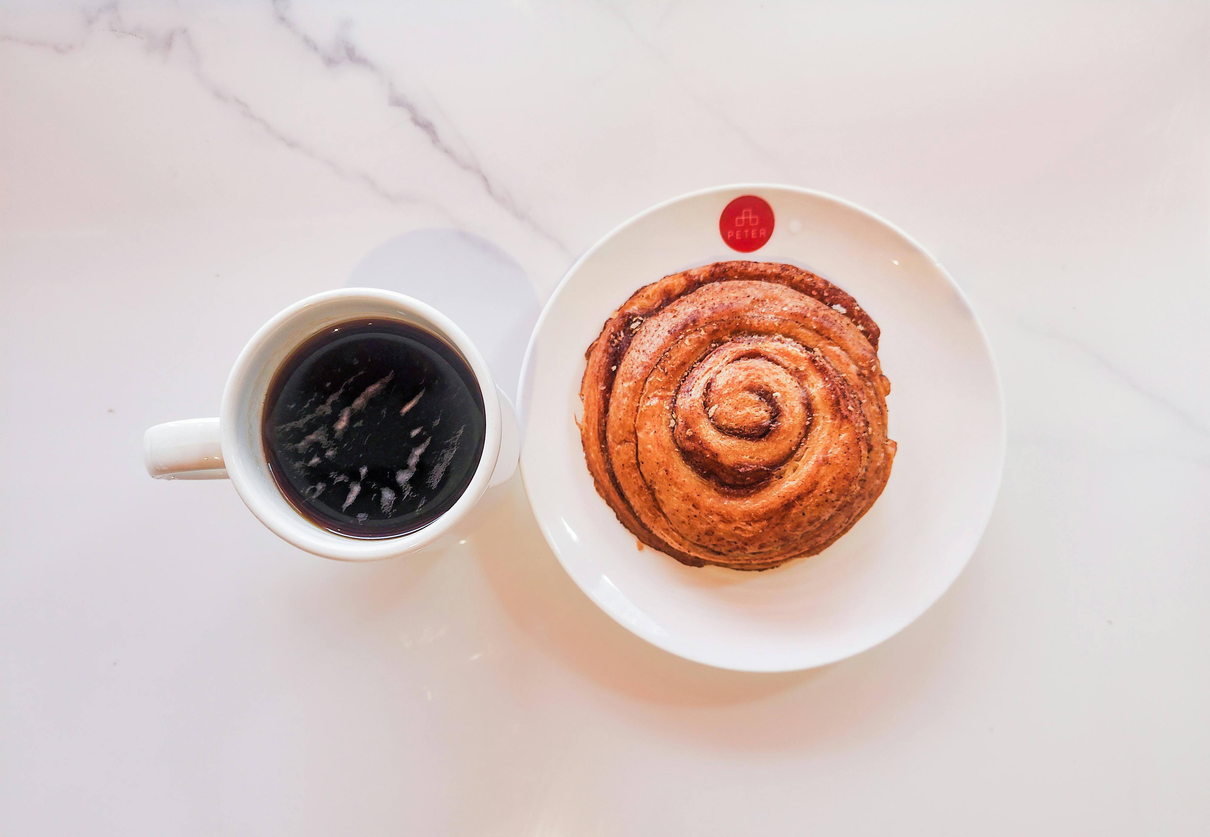 A top-view shot of black coffee with a cinnamon roll on a marble table surface.