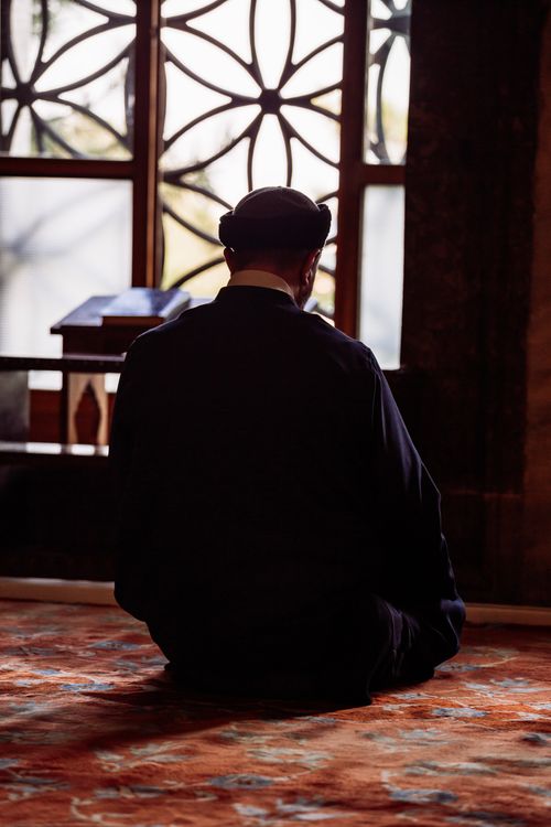 Free A man sitting and meditating on a patterned rug in a mosque with intricate window design. Stock Photo