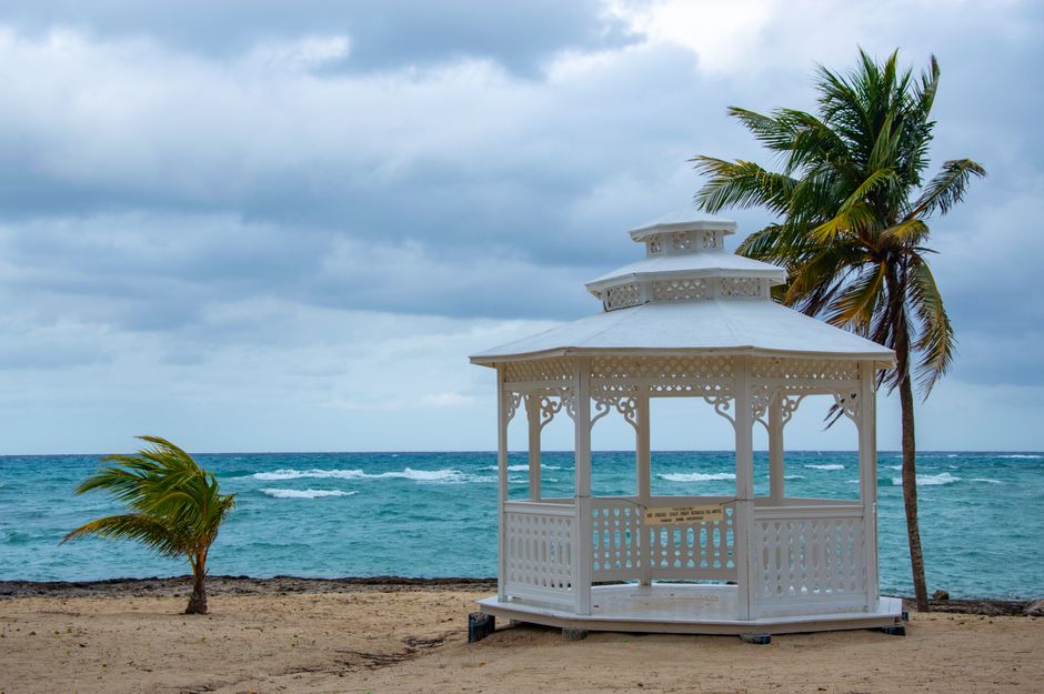 A serene tropical beach scene featuring a white gazebo, palm trees, and the ocean under a cloudy sky.