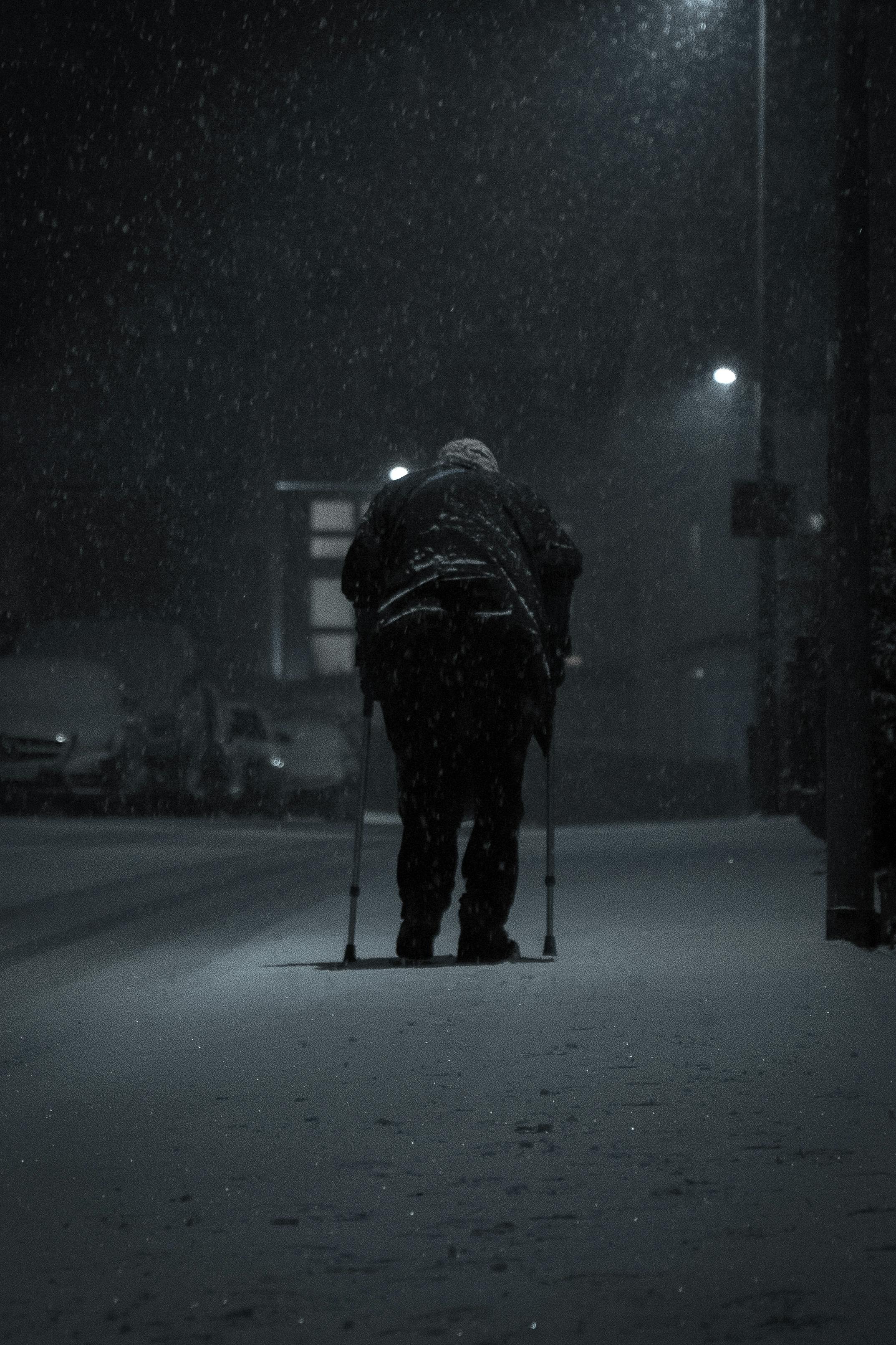 Elderly man struggles uphill in snowstorm at night using crutches.