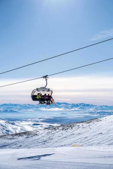 A ski lift carrying skiers over snowy mountains under a bright blue sky.