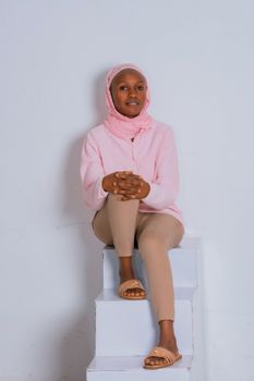 Young woman wearing hijab sits casually in a pastel outfit with a serene expression.