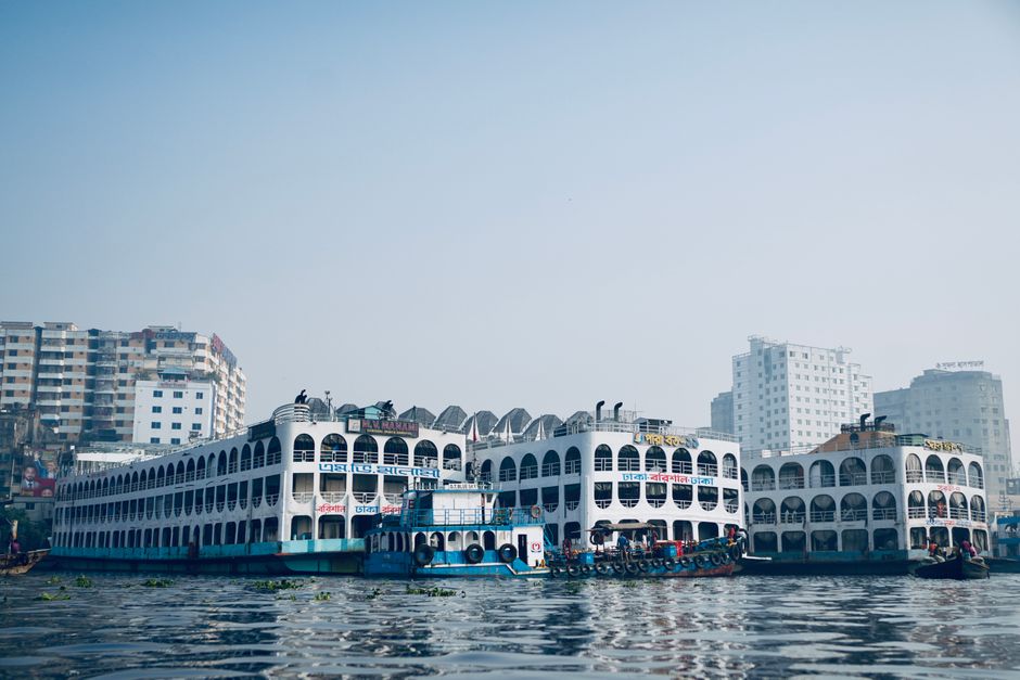 Passenger ferries docked at Sadarghat in Dhaka, Bangladesh, with urban buildings in the backdrop.