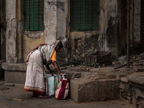 Free Elderly woman gathering bags on a rustic urban street with an aged building backdrop. Stock Photo