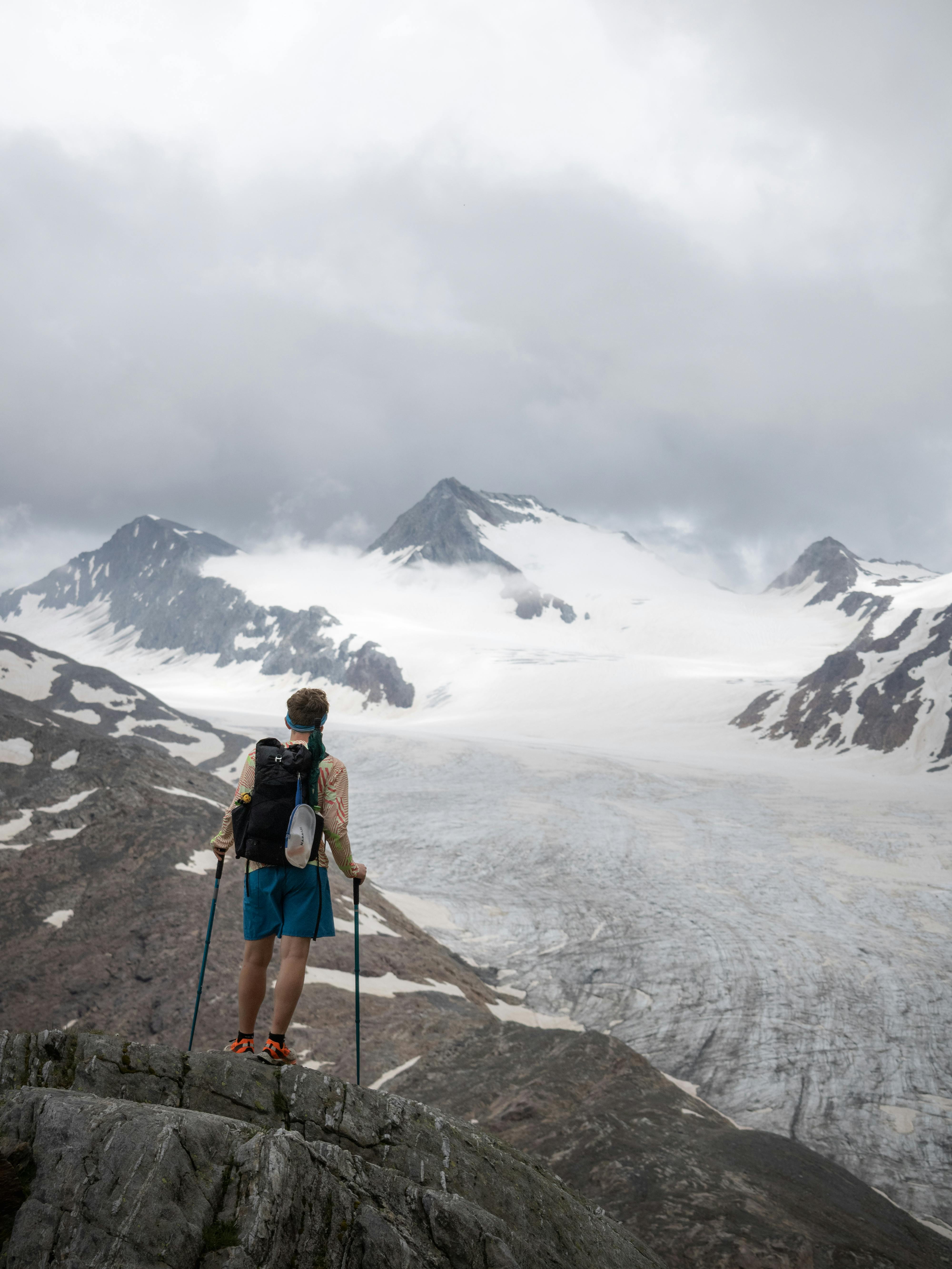 Hiker stands on a rocky peak overlooking majestic Obergurgl glaciers under a cloudy summer sky.