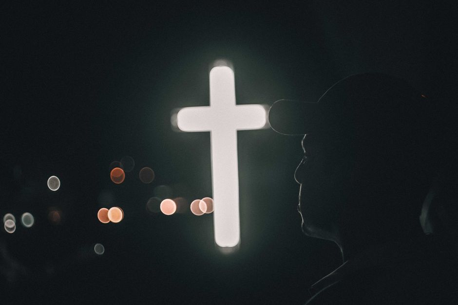 Silhouette of a man praying beside an illuminated cross at night, evoking spirituality and meditation.
