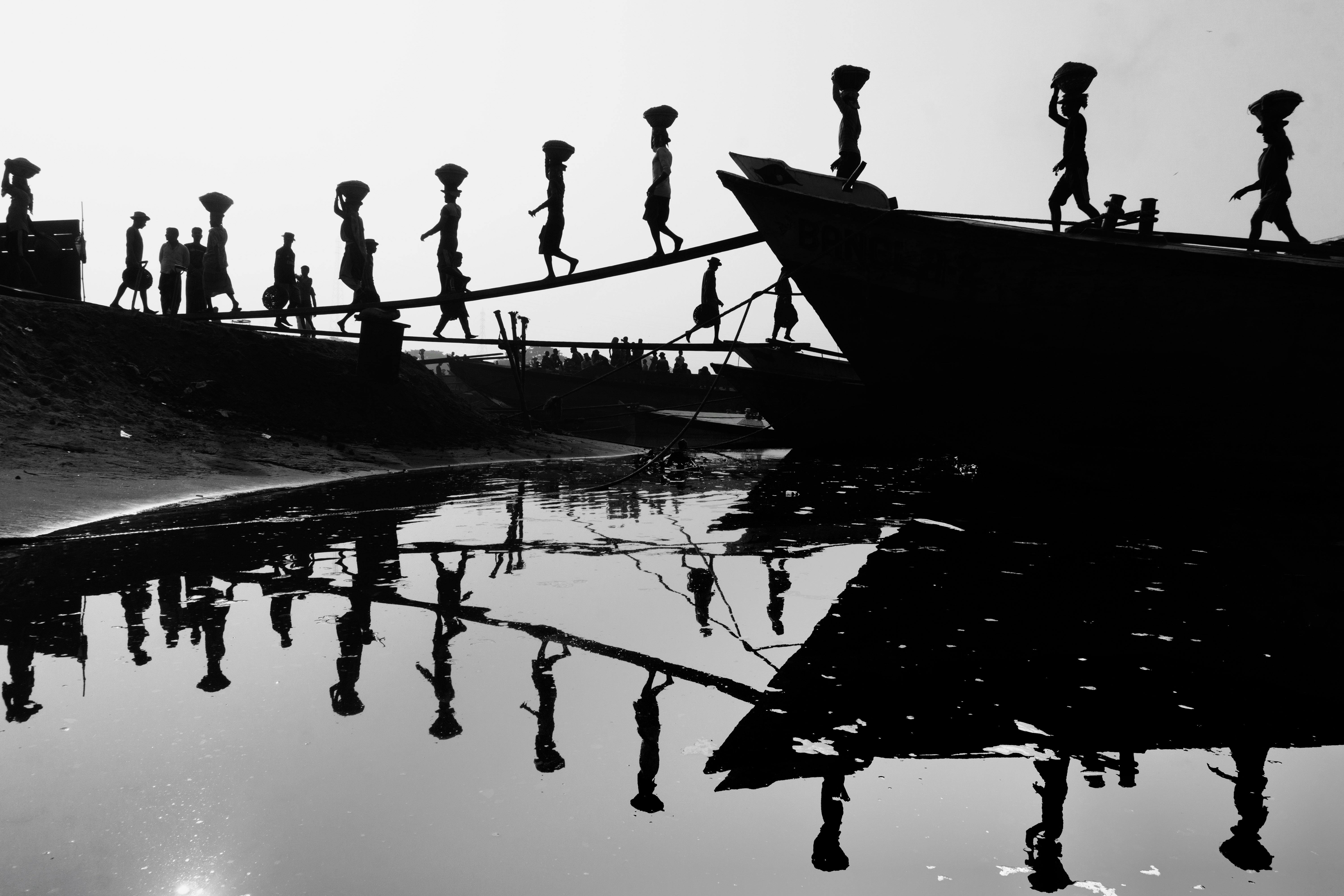Kostenlos Im Wasser von Bangladesch entladen Arbeiter, deren Silhouetten sich spiegeln, Fracht von einem Boot über eine Planke. Stock-Foto