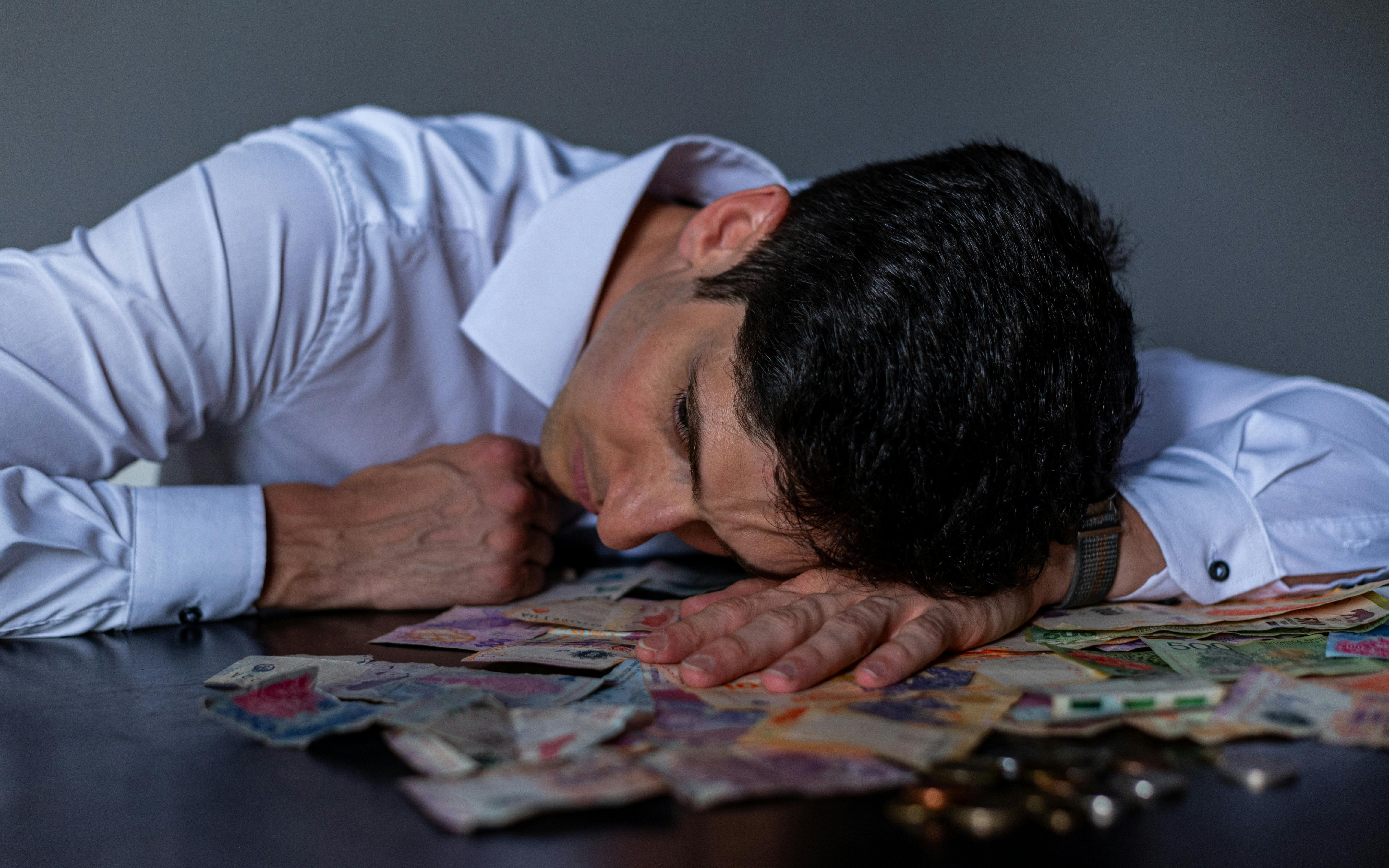 Man in stress resting on table covered with Argentine pesos symbolizing financial hardship.