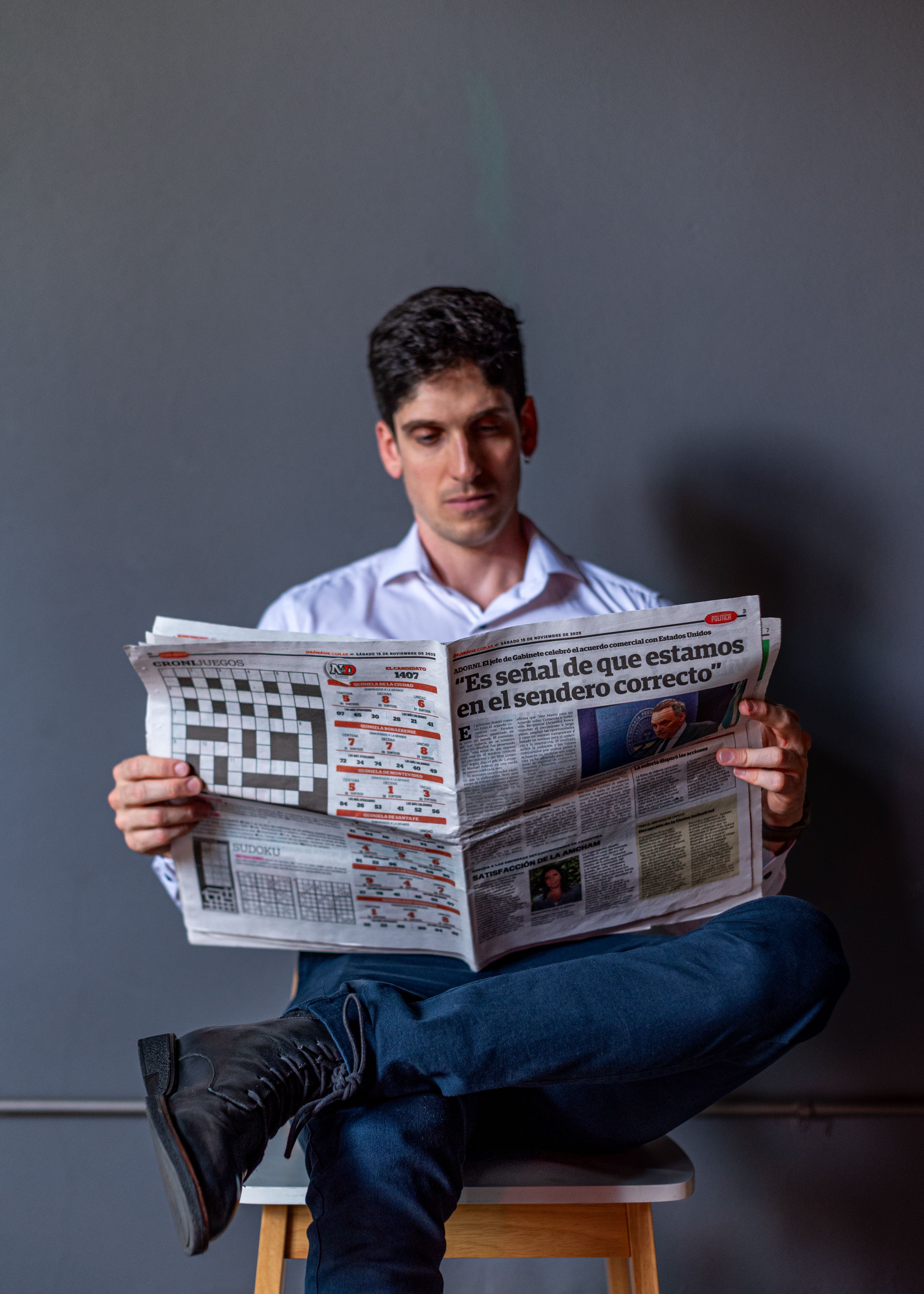 Focused man reading a newspaper in Spanish, sitting on a stool indoors.