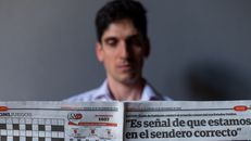 Man holding a Spanish newspaper with headline and date visible, indoors.