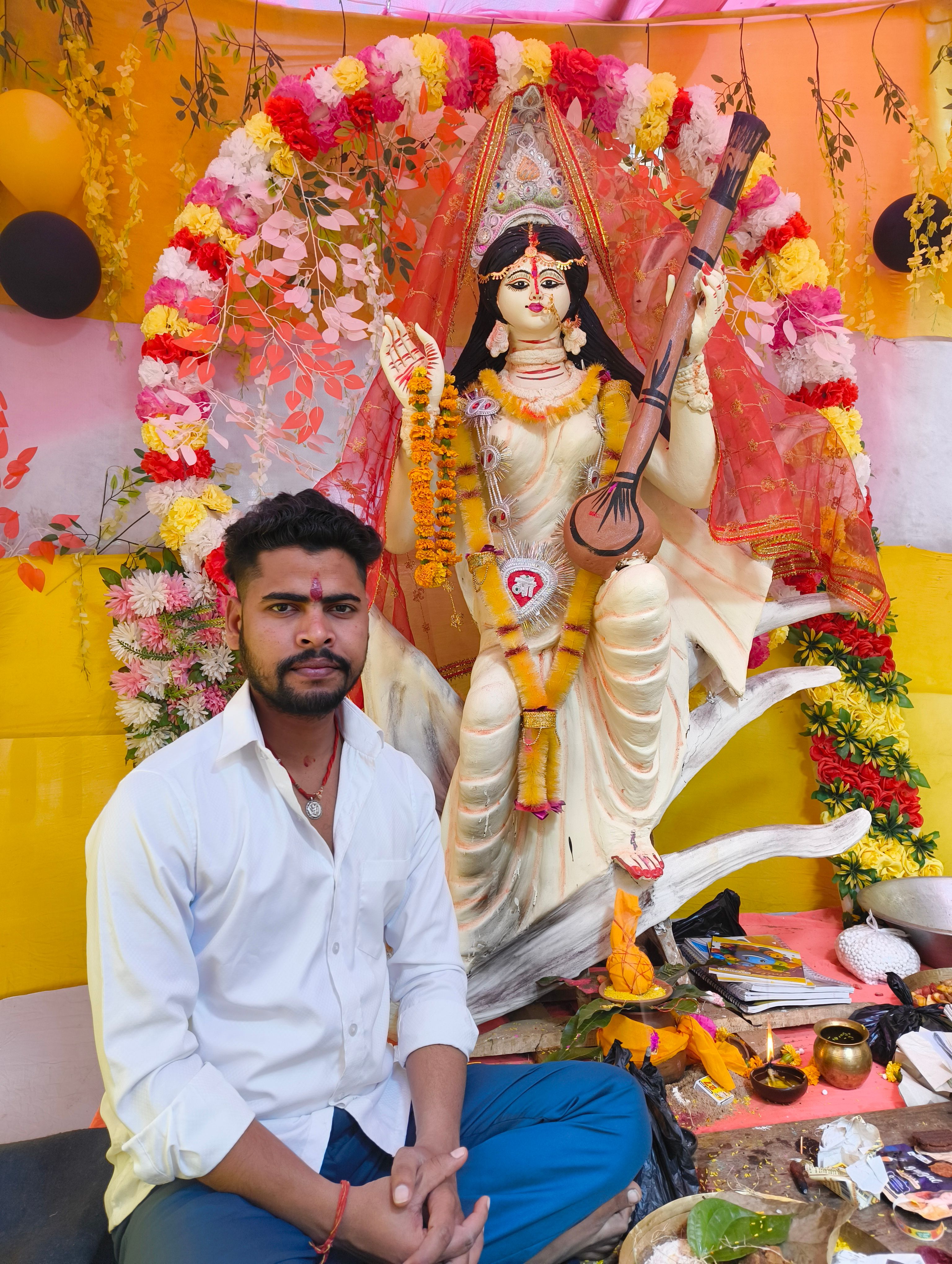 Devotee sitting near a decorated Idol of Goddess Saraswati with vibrant flowers.