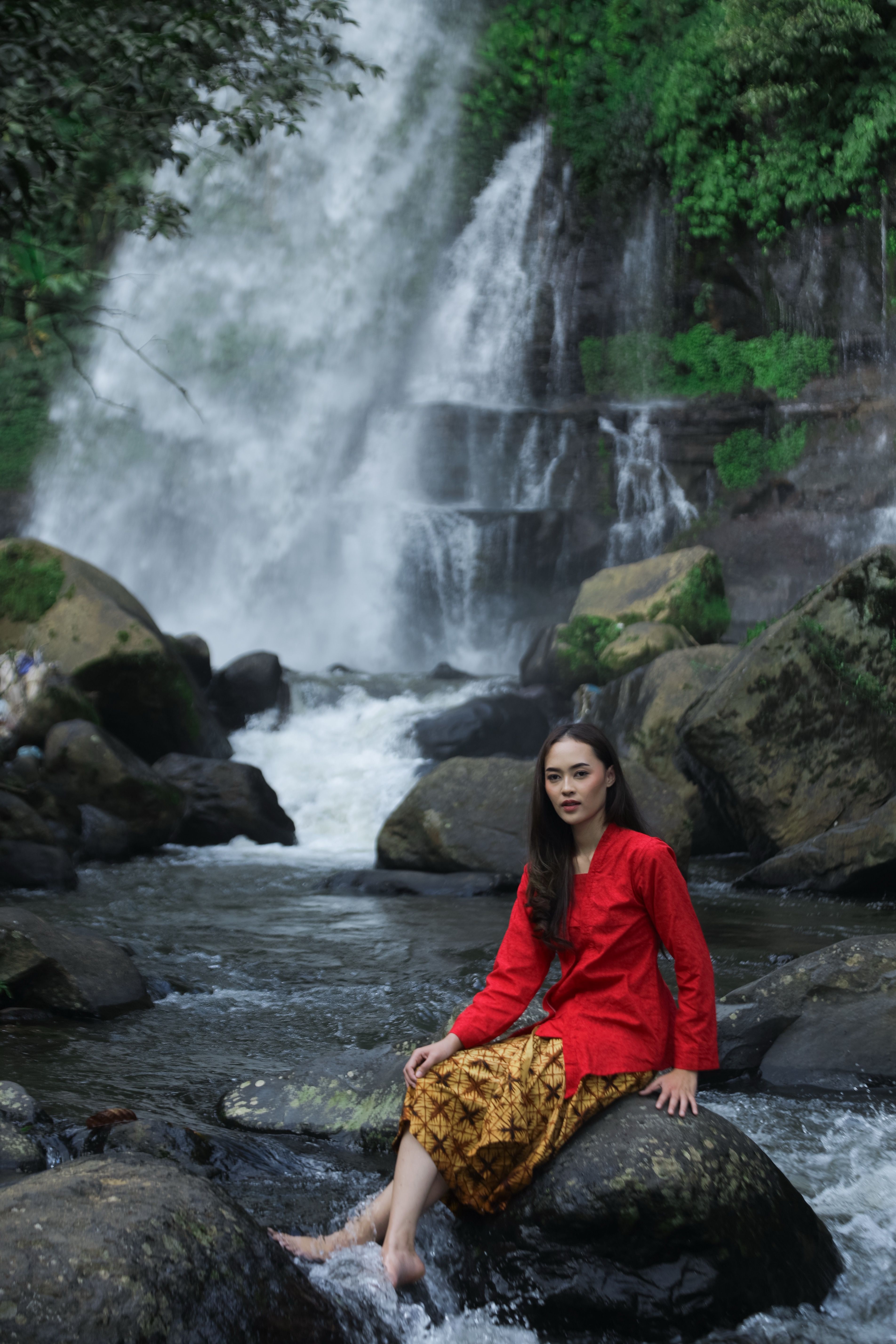 Young woman in traditional attire sitting by a waterfall, exuding natural beauty.