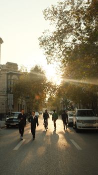 People walking and cycling in Baku, Azerbaijan during sunset on a tree-lined street.