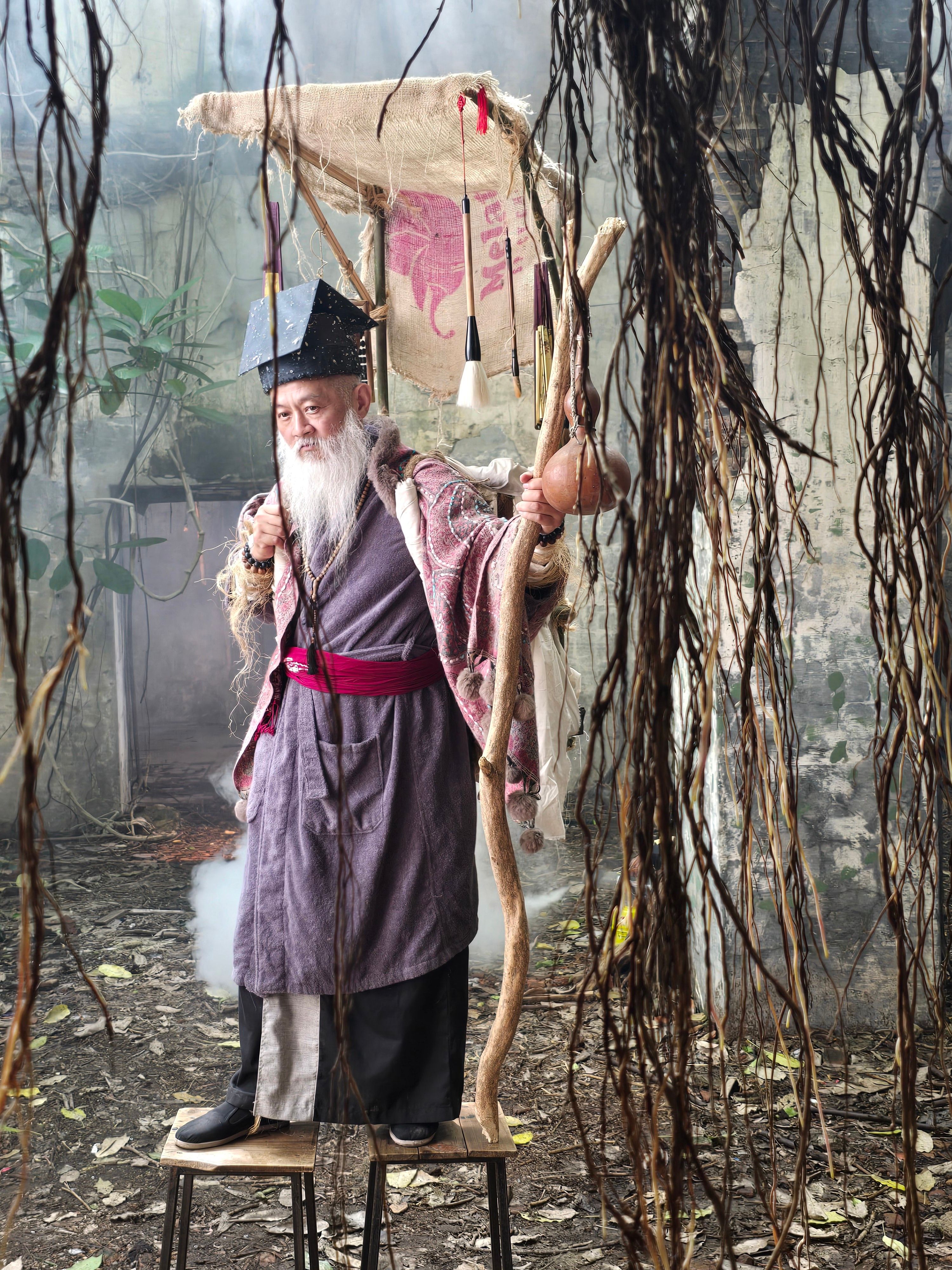 Elderly man in ancient robe and hat holding a wooden stick, standing outdoors.