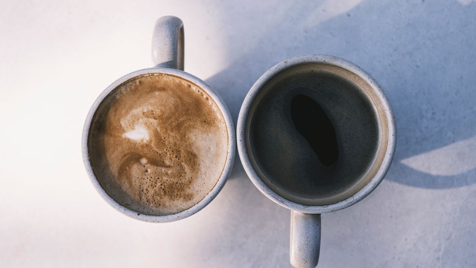 Two different coffees in grey cups casting shadows on a sunlit table