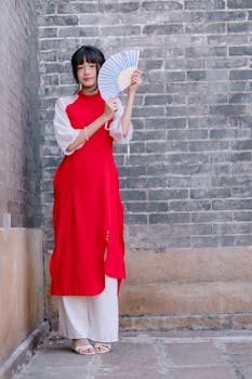 Portrait of a woman in a red dress holding a fan against a brick wall.