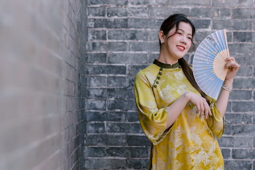 Smiling woman in a traditional yellow dress holds a decorative fan against a brick wall backdrop.