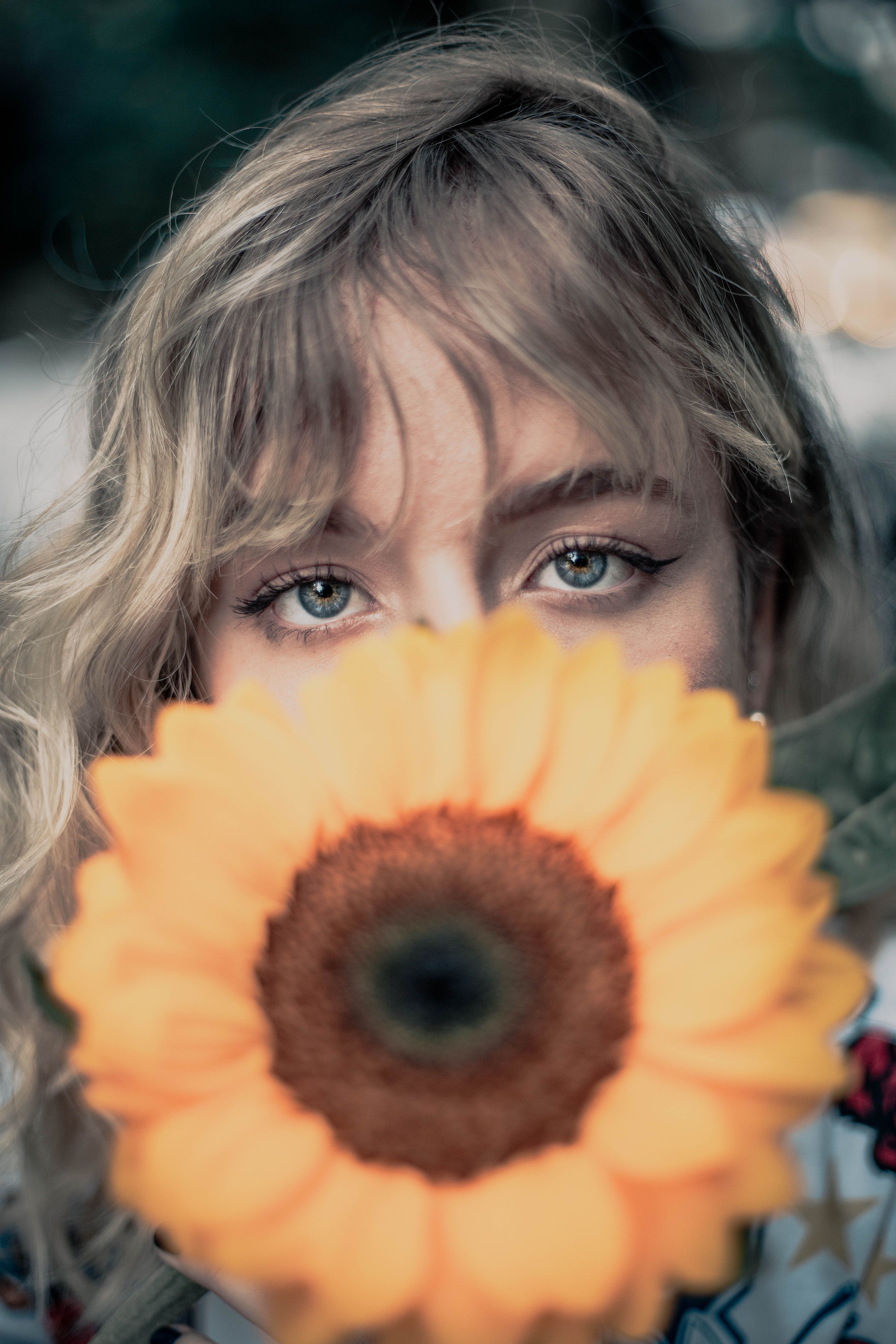 Blonde woman with striking eyes gazes through a sunflower in a vibrant São Paulo setting.