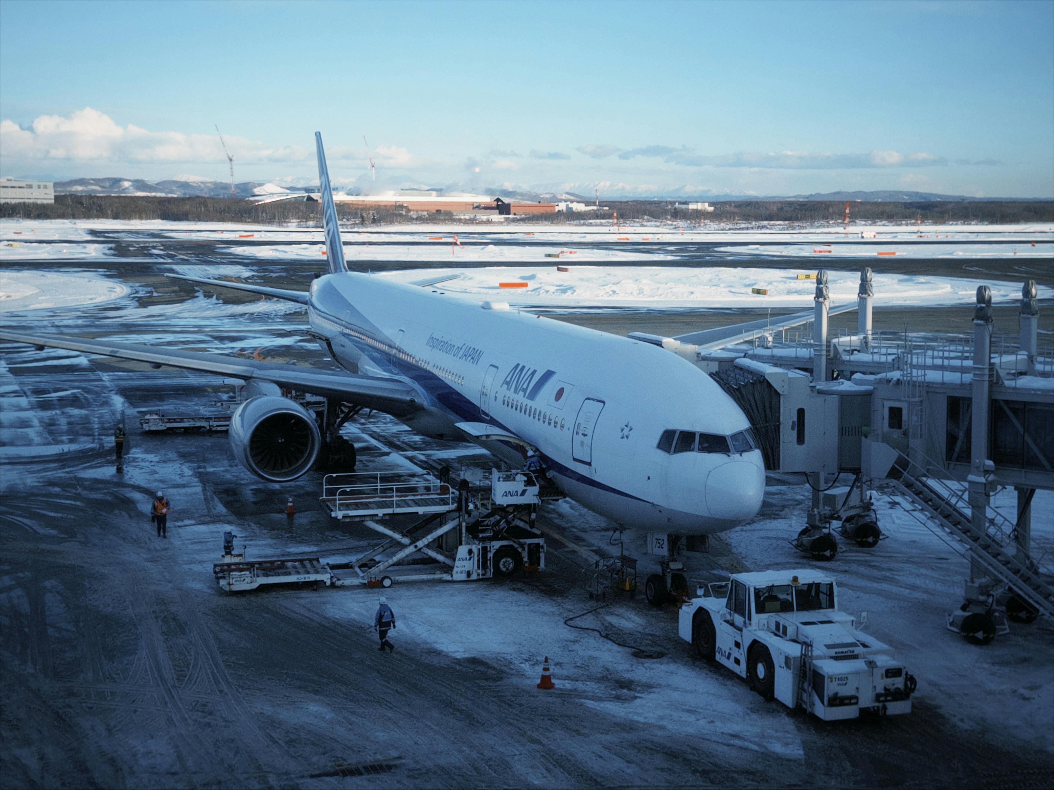An airplane parked at New Chitose Airport in winter, surrounded by snow and ground crew.