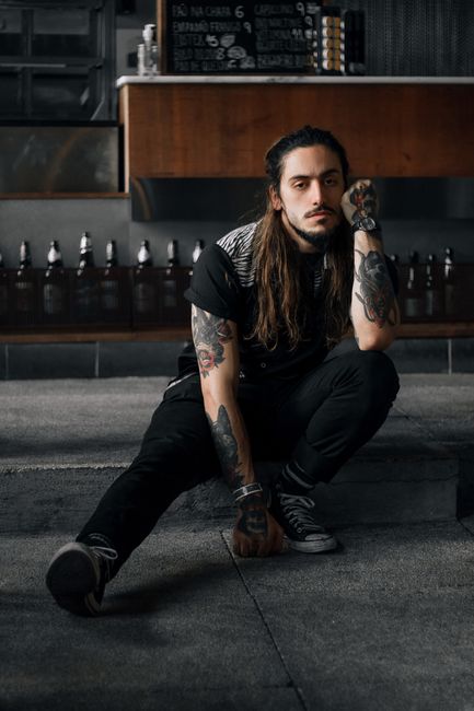 A tattooed man sitting indoors, showcasing fashion and tattoos in a casual pose.