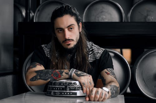 Man With Long Hair And Tattoos Sitting On A Chair Behind A Table