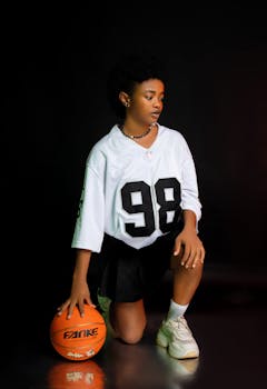 Young basketball player in jersey with ball, posing against black backdrop.