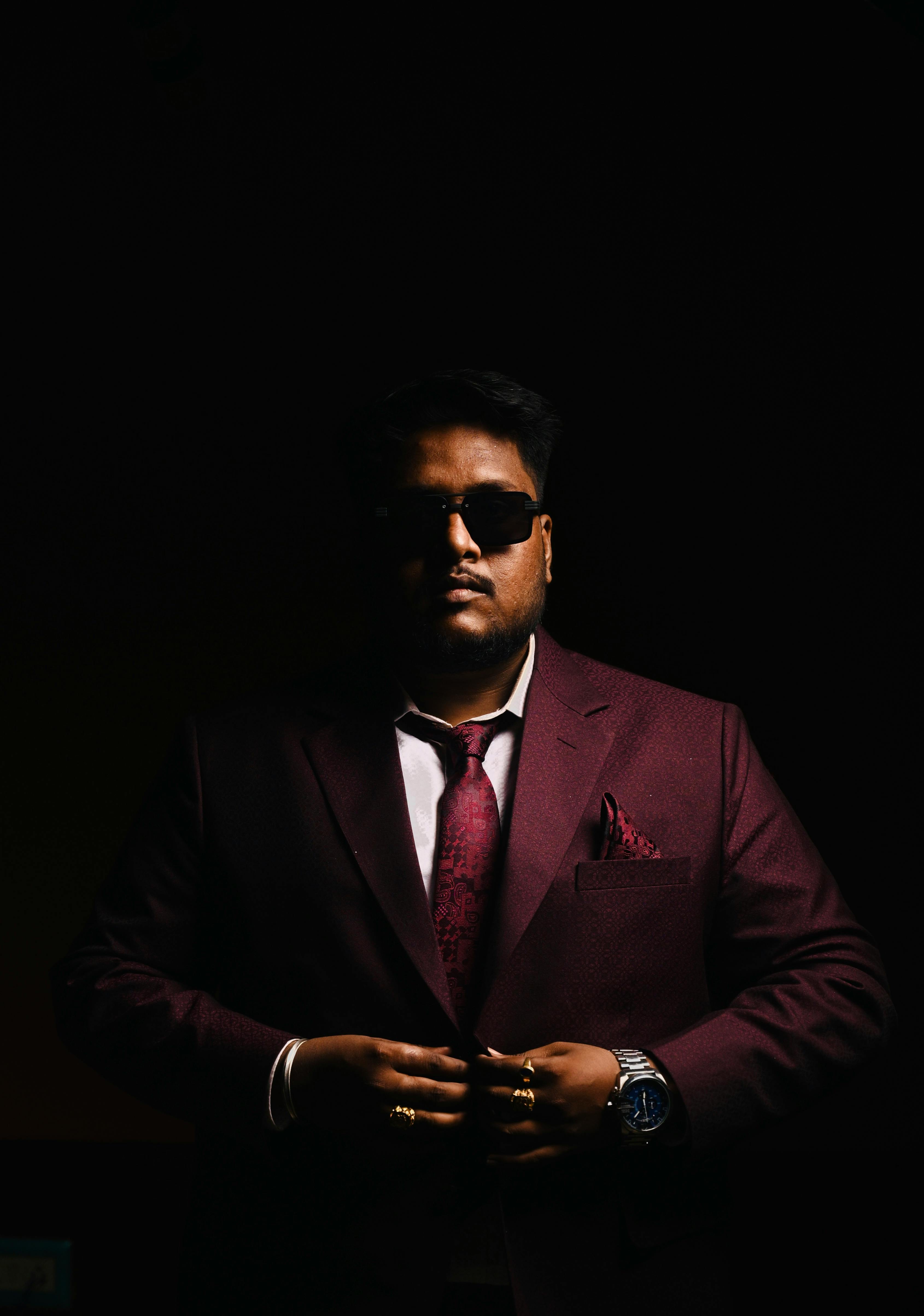 A stylish man in a burgundy suit and sunglasses exudes confidence against a dark backdrop.