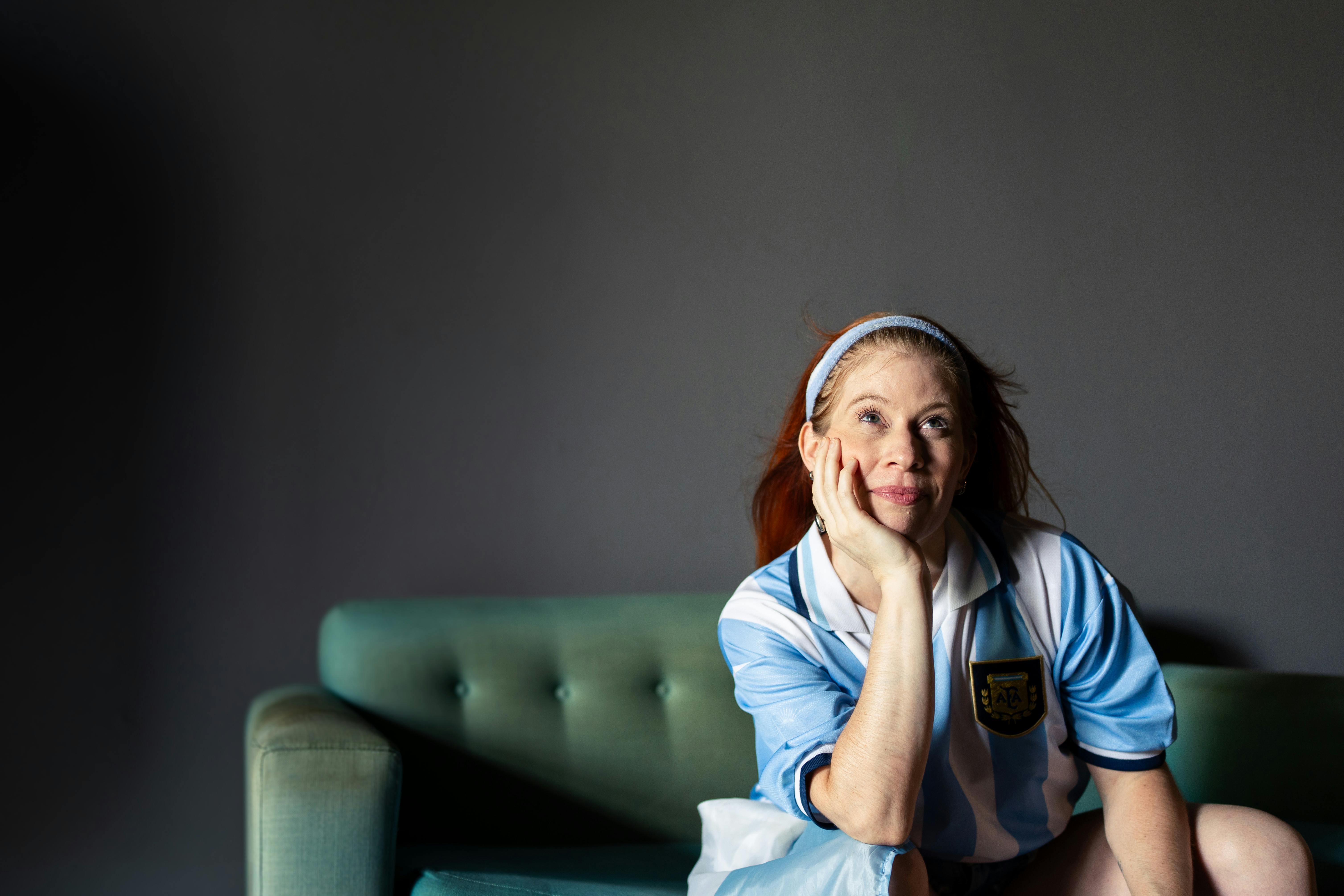 A woman happily seated on a green couch wearing an Argentina football jersey, indoors.